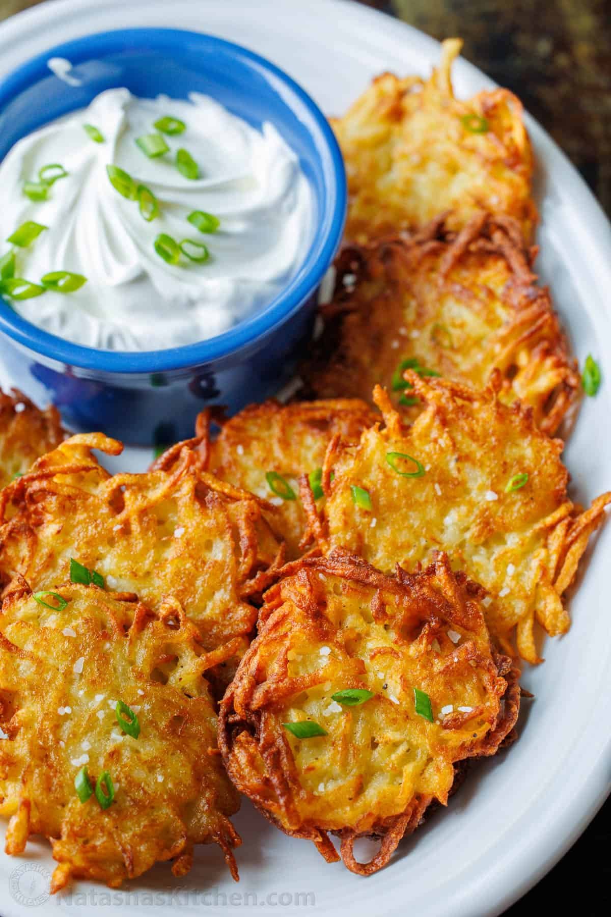 This latke recipe creates the crispiest potato fritters. Sprinkled with salt and dolloped with sour cream, these potato latkes are total comfort food. Latkes fried and topped with chives. On a plate with sour cream