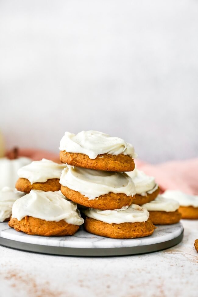 pumpkin cookies with cream cheese frosting stacked on marble tray.