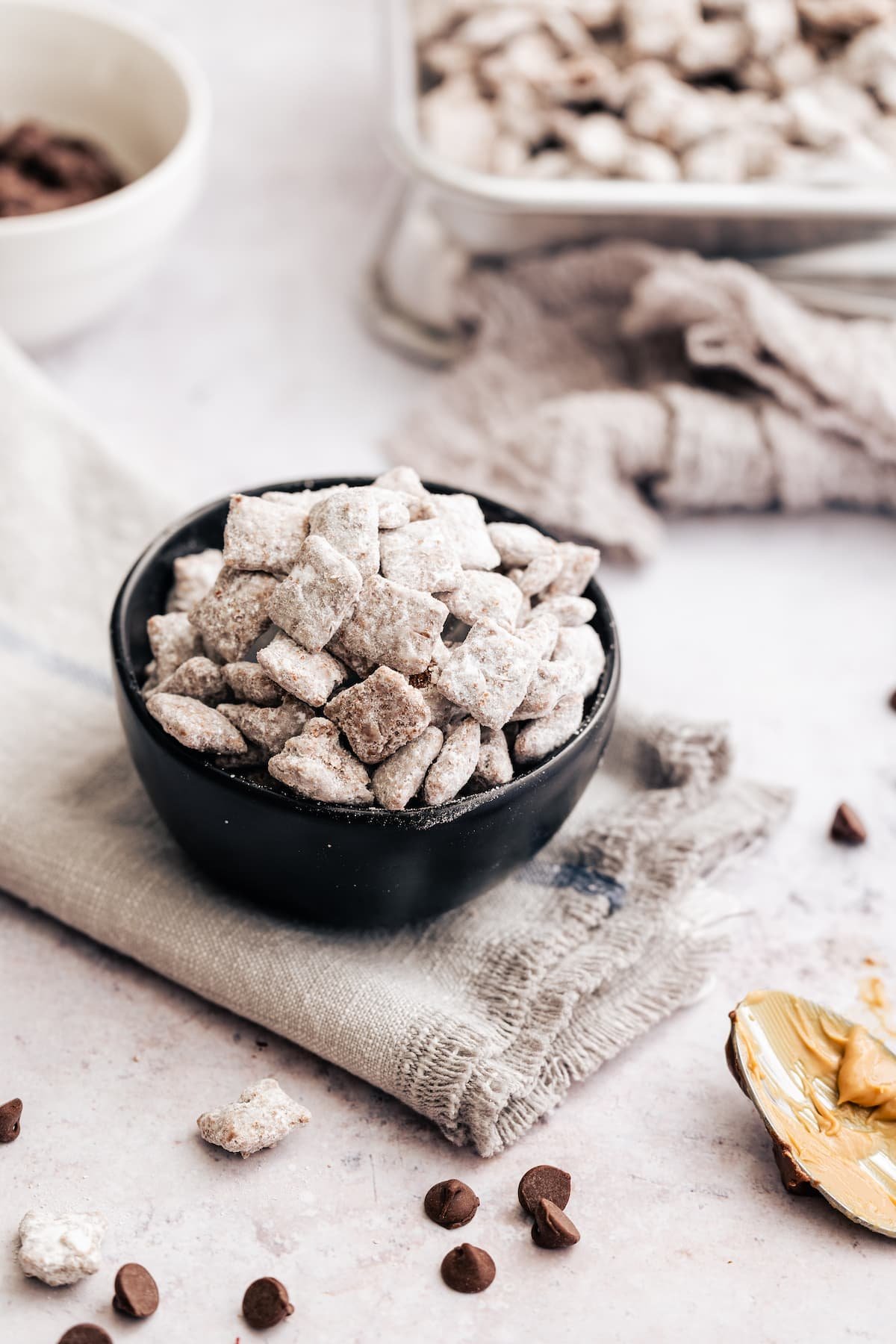 Homemade Puppy Chow is easy to make and SO addicting. This recipe for chocolate peanut butter-covered Chex cereal is a perfect party snack! A bowl of puppy chow on a grey cloth, surrounded by scattered puppy chow and a spoon covered with peanut butter.