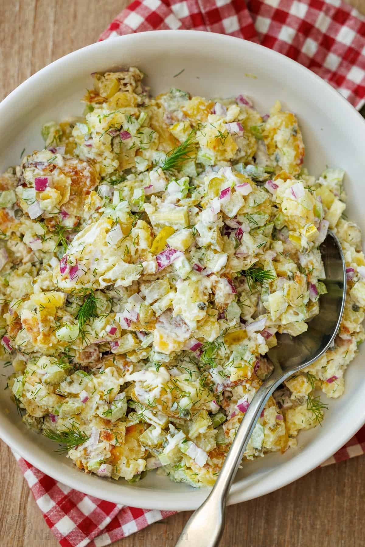 Smashed Potato Salad has a delicious twist with crispy smashed potatoes, fresh veggies, herbs, and the best 2-ingredient dressing. So good! Smashed Potato Salad in a white bowl with a serving spoon, sitting on a red checkered towel.