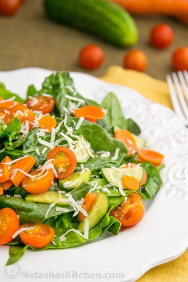 A plate of spinach salad with tomatoes and cucumbers in the background and a fork beside the plate