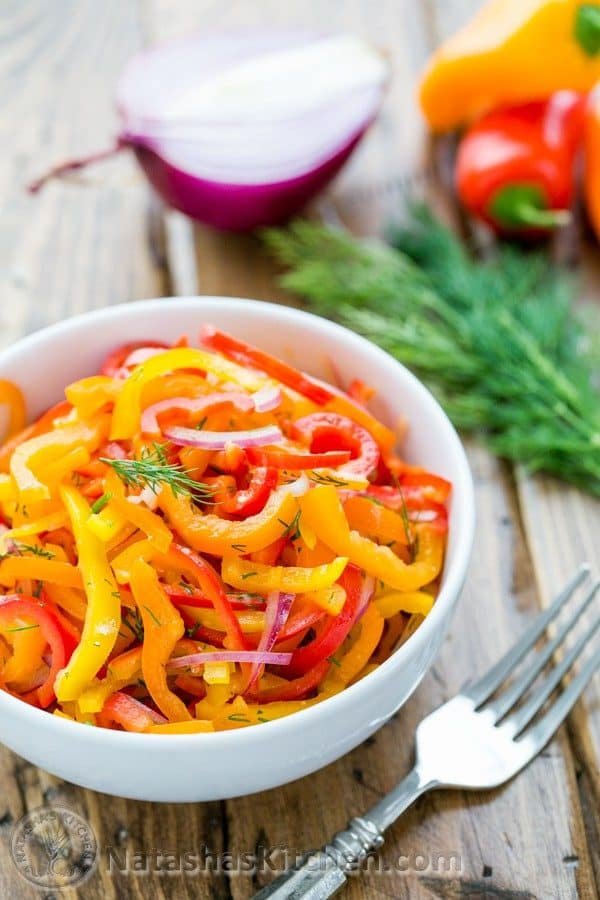 A bowl of zesty mini bell pepper salad with a fork, dill, mini peppers and purple onion beside it