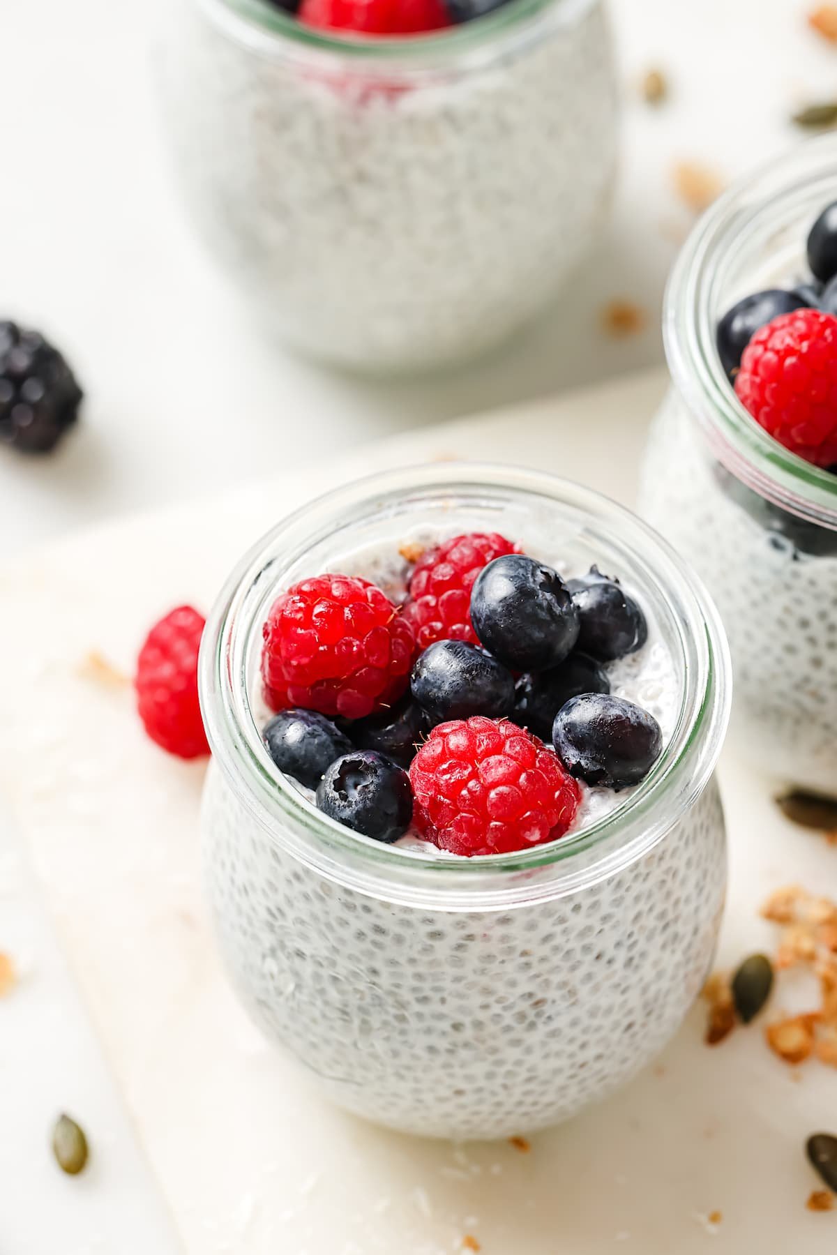 chia pudding being stirred in small glass jar on marble board with tea towel and berries. 