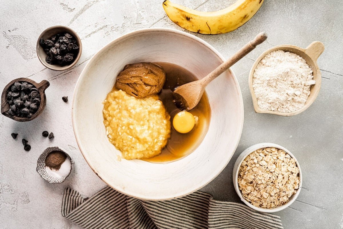 mashed bananas, nut butter, maple syrup, egg, and vanilla in mixing bowl with wooden spoon to make breakfast cookies.