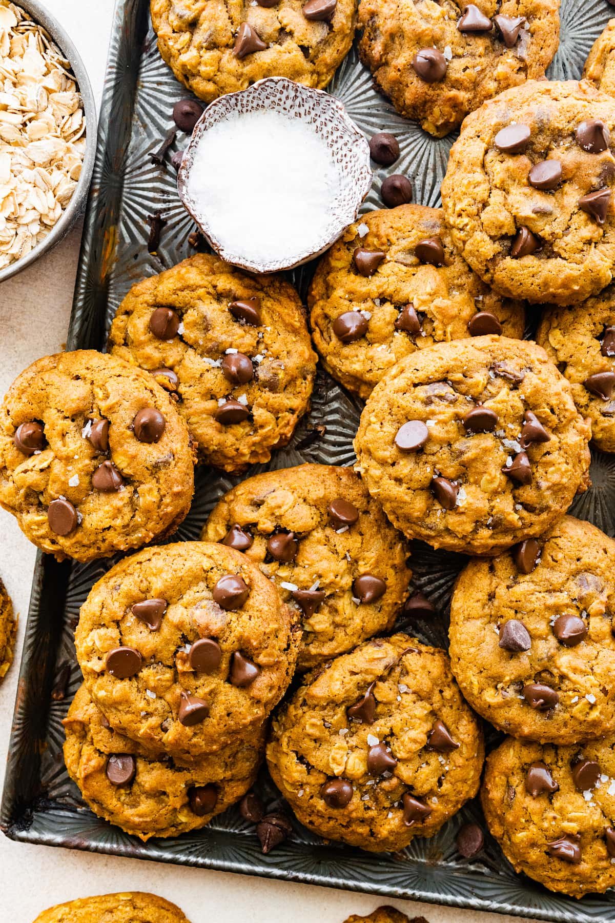 pumpkin oatmeal chocolate chip cookies on baking sheet with chocolate chips and little bowl of sea salt.