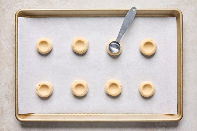 thumbprint cookie dough pressed into thumbprints with a teaspoon on a baking sheet lined with parchment paper.