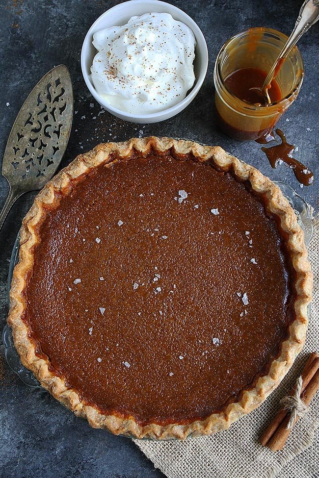 Salted Caramel Pumpkin Pie Is The BEST Pumpkin Pie Recipe. This Pumpkin Pie Will Be Everyone's Favorite Thanksgiving Dessert! #pumpkin #pumpkinpie #pie #pierecipe #Thanksgiving #Christmas #holidays #baking #Thanksgivingrecipe salted caramel pumpkin pie in pie plate with a bowl of whipped cream and jar of salted caramel sauce.
