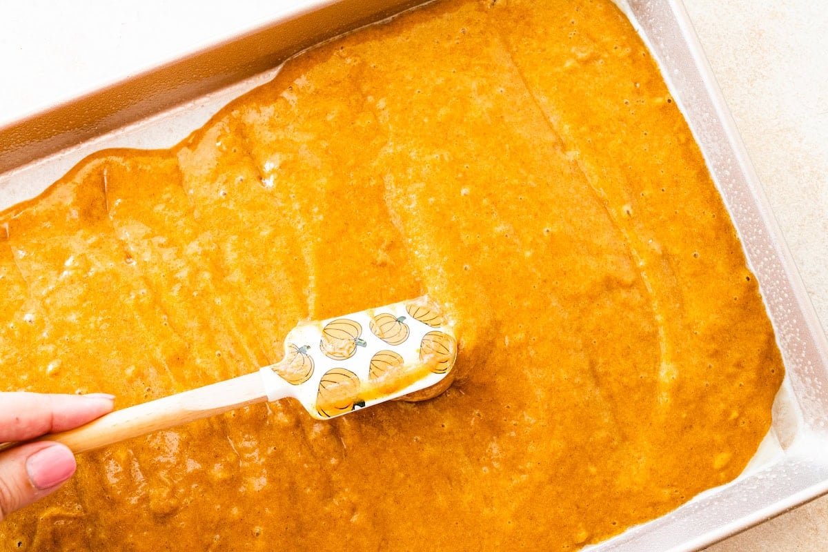 spreading pumpkin roll cake batter onto sheet pan with spatula.