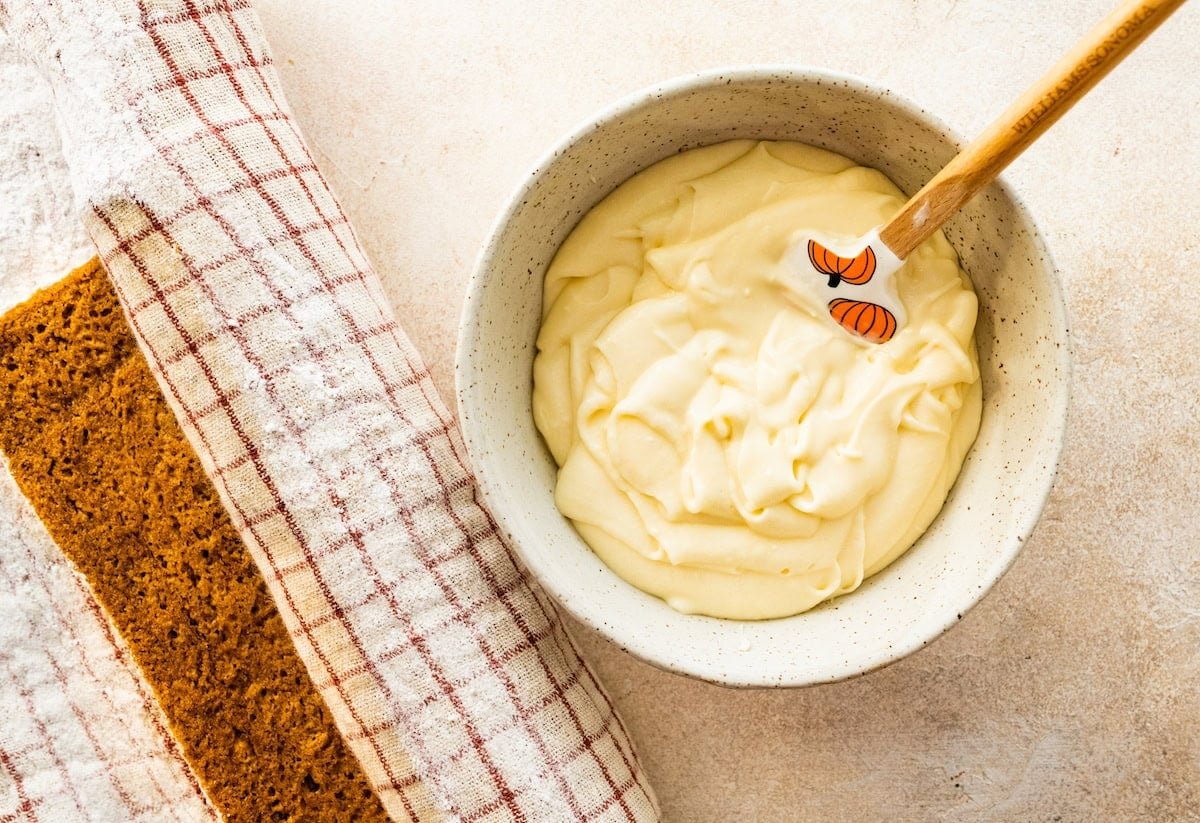 cream cheese frosting in bowl with spatula with pumpkin roll in kitchen towel.