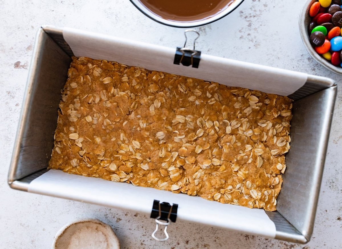 peanut butter oat mixture pressed in loaf pan lined with parchment paper to make no bake monster cookie bars.