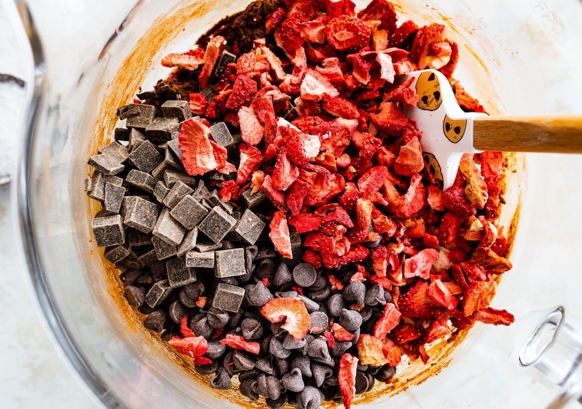 chocolate chips, chocolate chunks, and freeze dried strawberries being stirred into cookie dough with spatula.