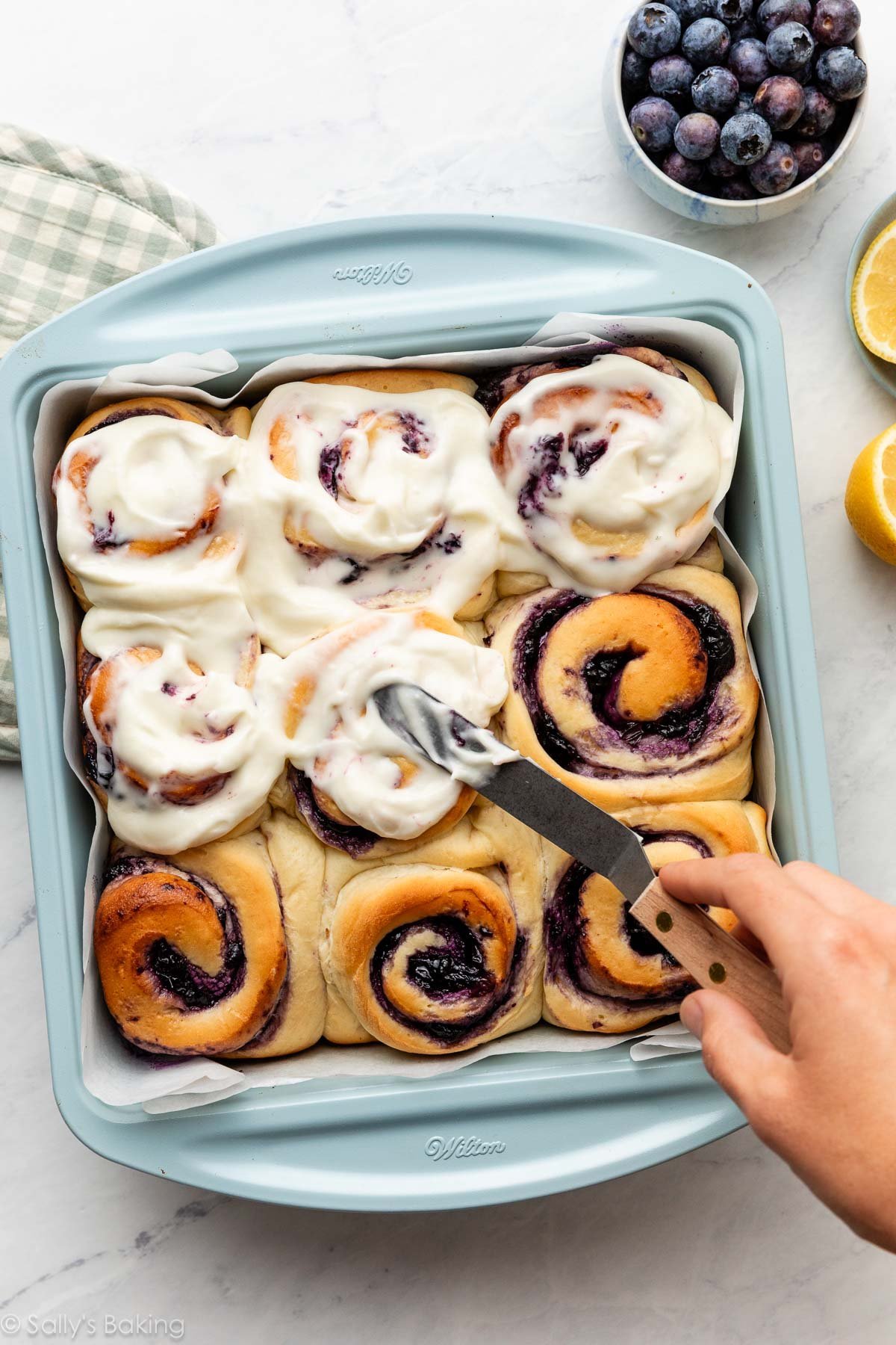 spreading frosting on blueberry rolls in blue pan.