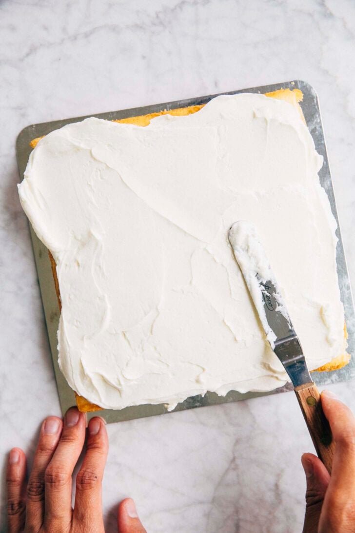 photo of Michelle from Hummingbird High covering the first white almond cake layer with chantilly cream cheese frosting