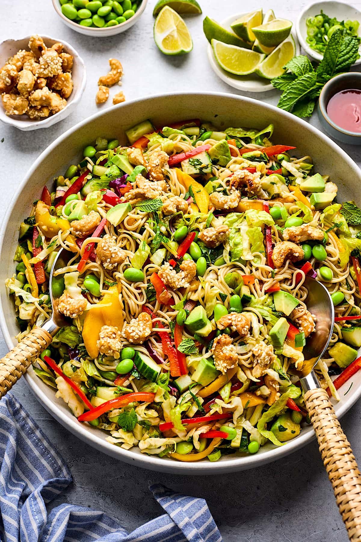 sesame noodle salad with mango, red pepper, edamame, avocado, herbs, and honey sesame cashews in sesame ginger dressing in bowl with salad servers.