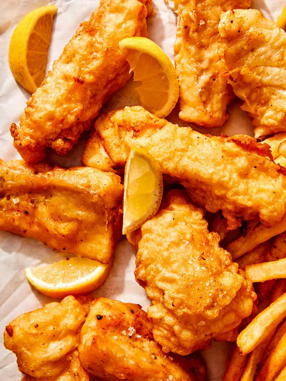 Close up on a bunch of beer battered fish on parchment with lemons and fries peeking in.