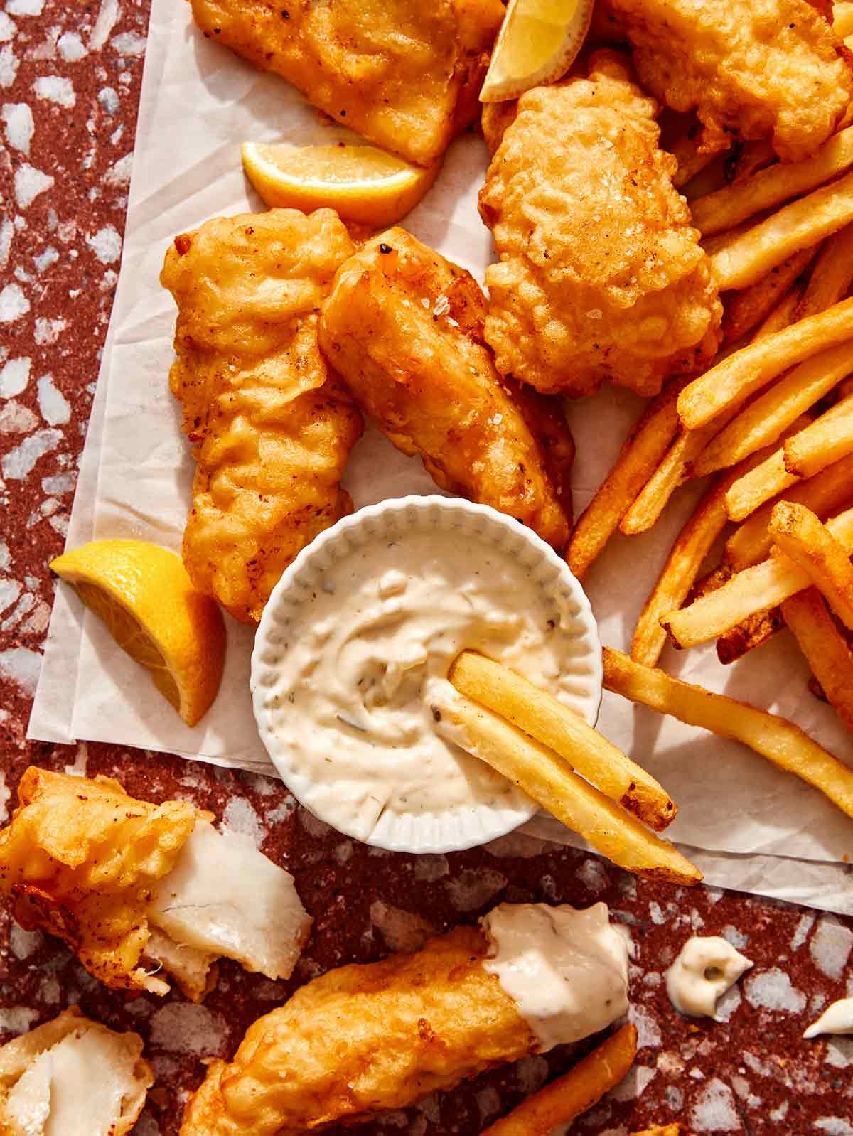 Beer battered fish on parchment with french fries and tartar sauce with some fries dipping in the sauce.