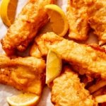 Close up on a bunch of beer battered fish on parchment with lemons and fries peeking in.