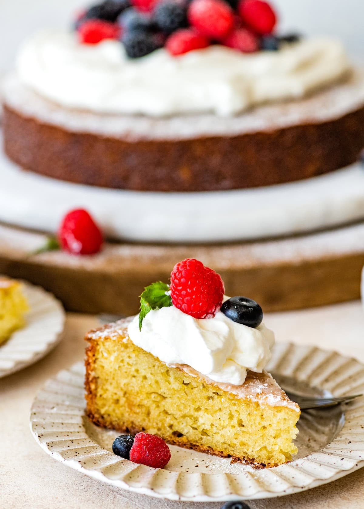 Slice of yogurt cake on plate with whipped cream and berries with cake in the background.