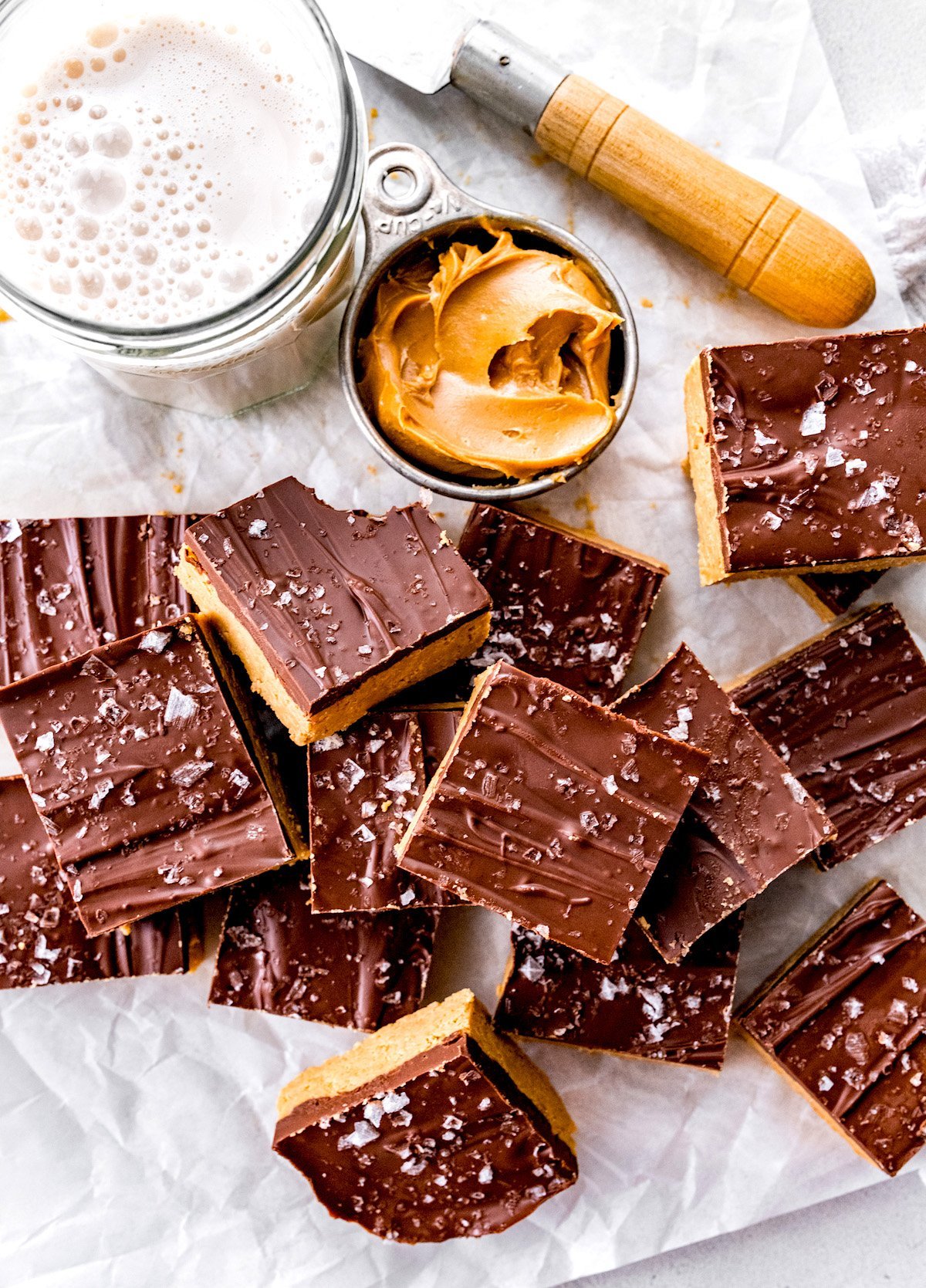 no bake peanut butter bars on parchment paper with a cup of peanut butter, glass of milk, and knife.