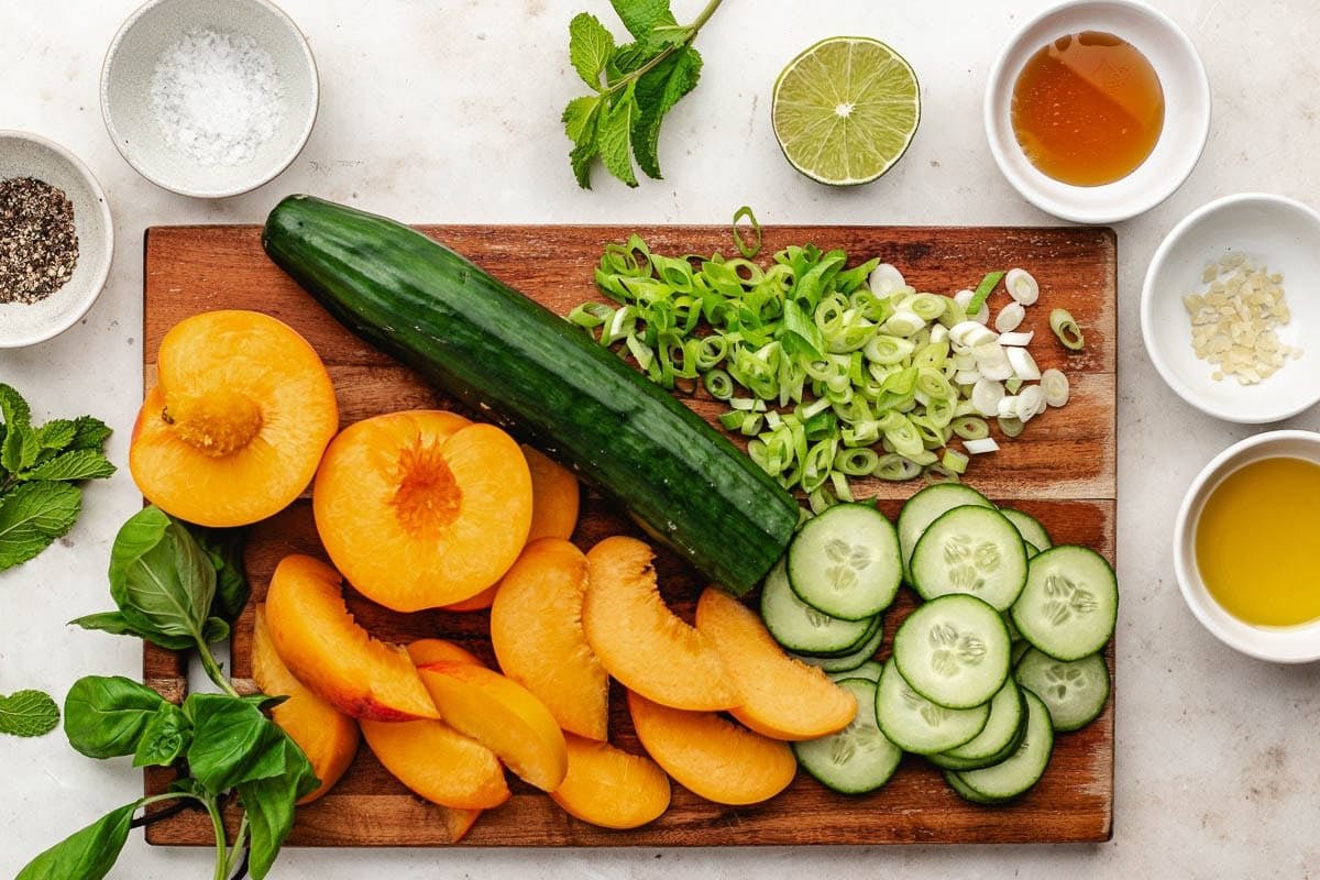 peaches, cucumber, green onion, herbs on wood cutting board and ingredients in bowls to make peach cucumber salad. 