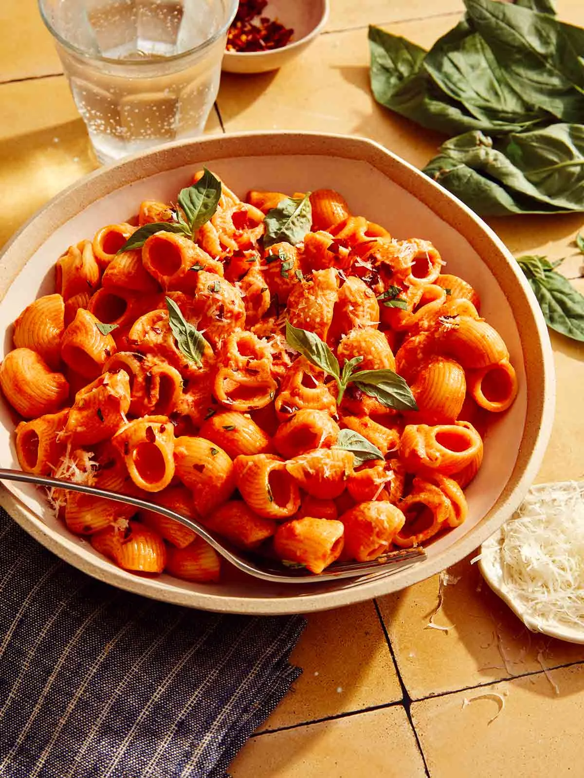 Spicy rigatoni in a bowl with a fork on the side and some cheese and basil on the table.