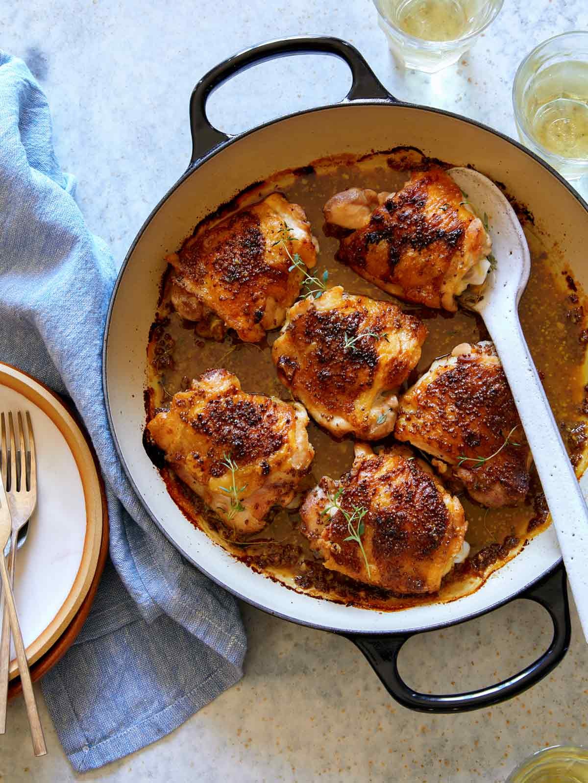 Oven baked chicken thighs in a skillet with glasses of wine on the side.