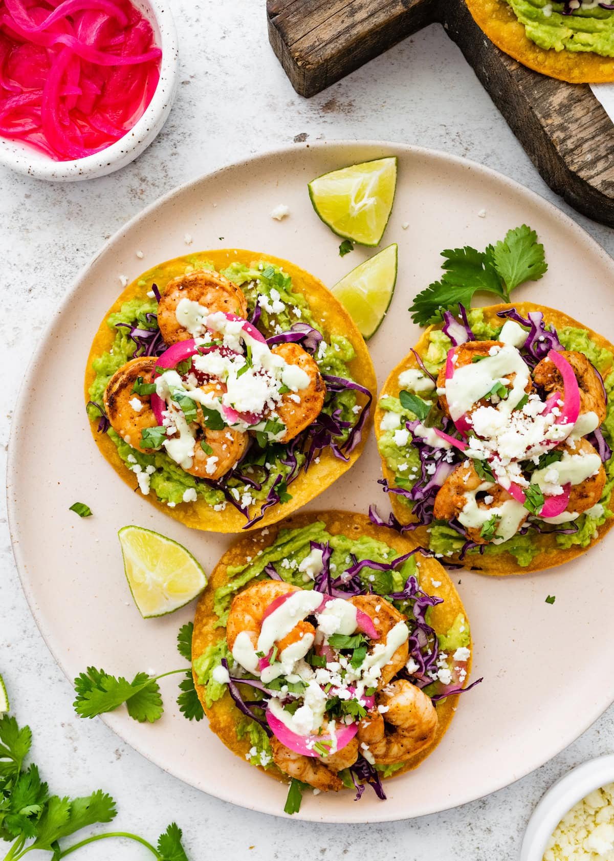 three shrimp tostadas on plate with pickled red onions, lime wedges, and cilantro.