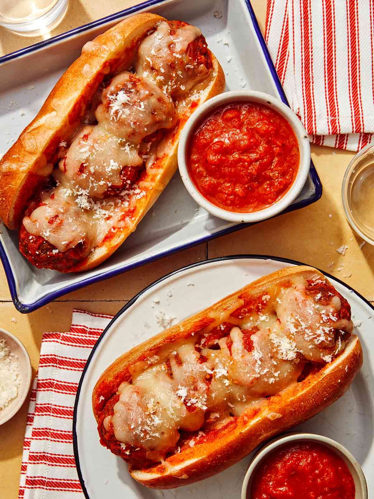 Two meatball subs on plates with marinara sauce on the side.