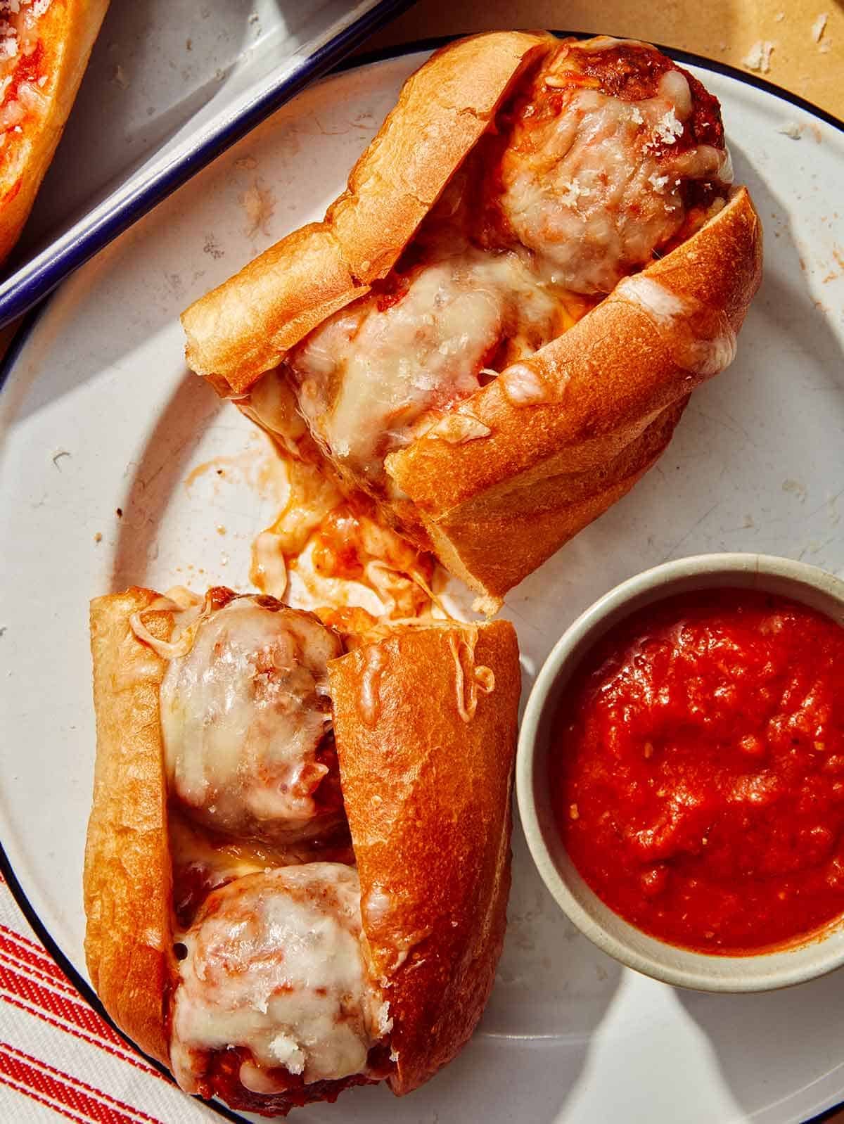 A meatball sub on a plate cut in half with marinara sauce on the side.