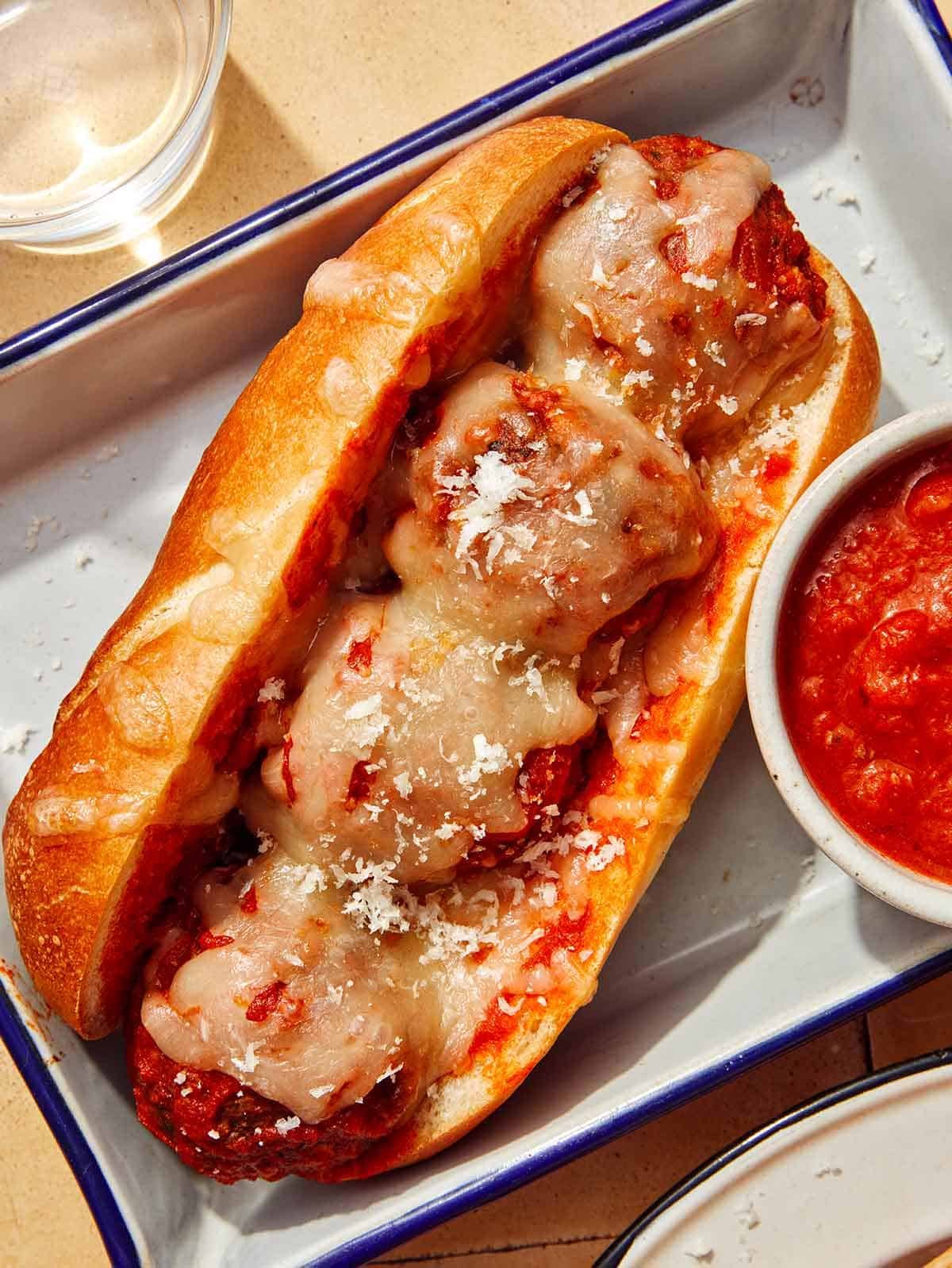 Close up on a meatball sub with water in the background and a side of marinara sauce.