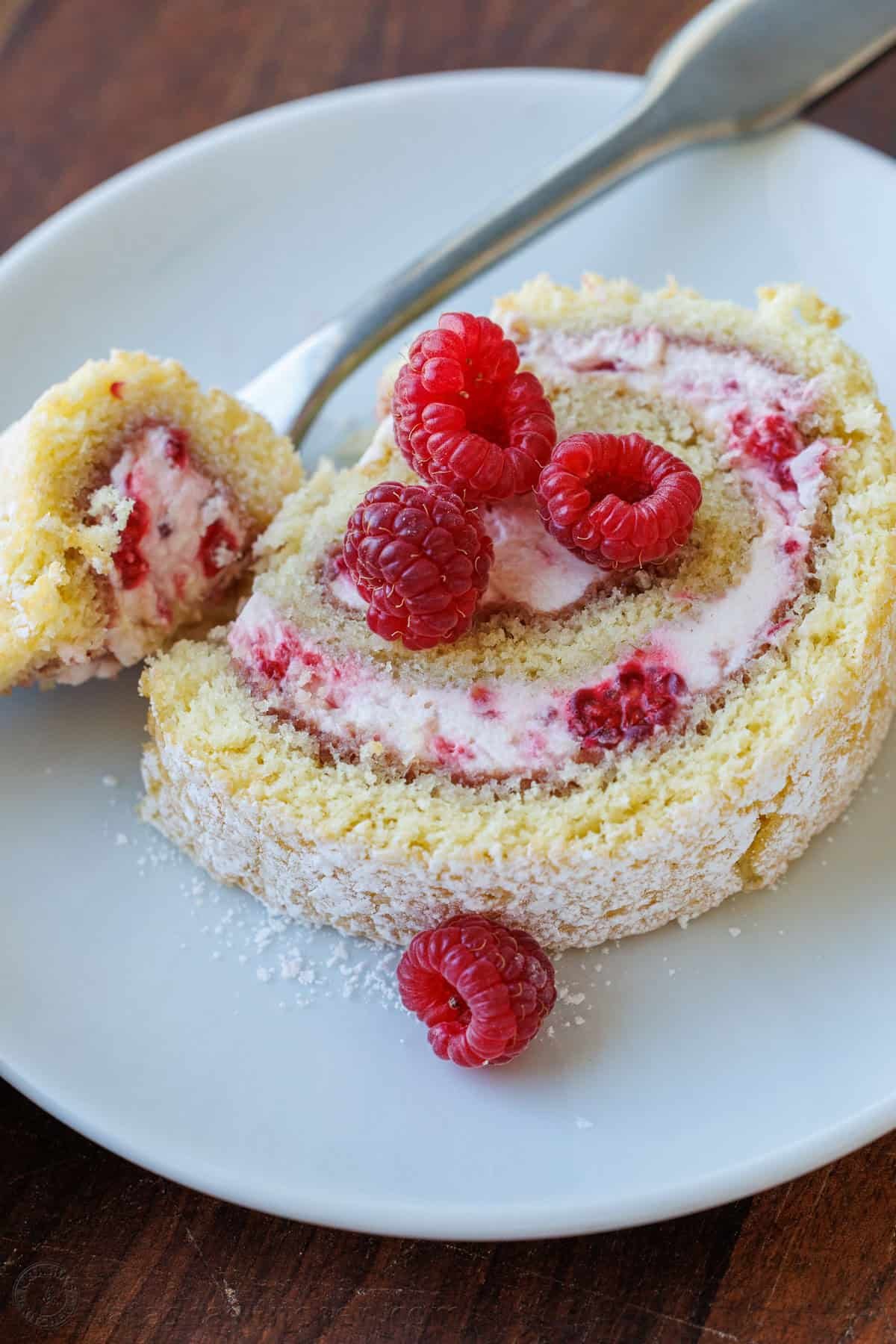 Raspberry Swiss Roll Cake wraps moist sponge cake around a raspberry cream filling. This Raspberry Cake Roll is gorgeous and simple to make. A slice of raspberry cake roll on a white plate with a fork holding a bite.