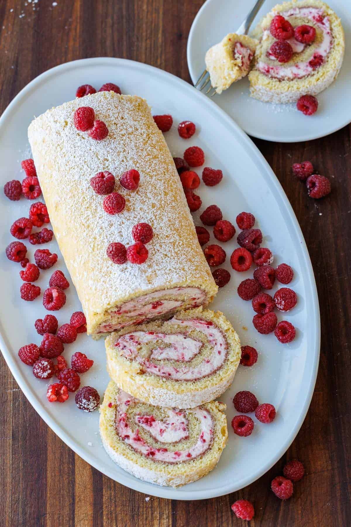 Raspberry Swiss Roll Cake wraps moist sponge cake around a sweet and creamy mascarpone filling. Sounds difficult, but it couldn't be easier! Swiss Roll cake recipe with raspberries on a white plate and two slices cut out.