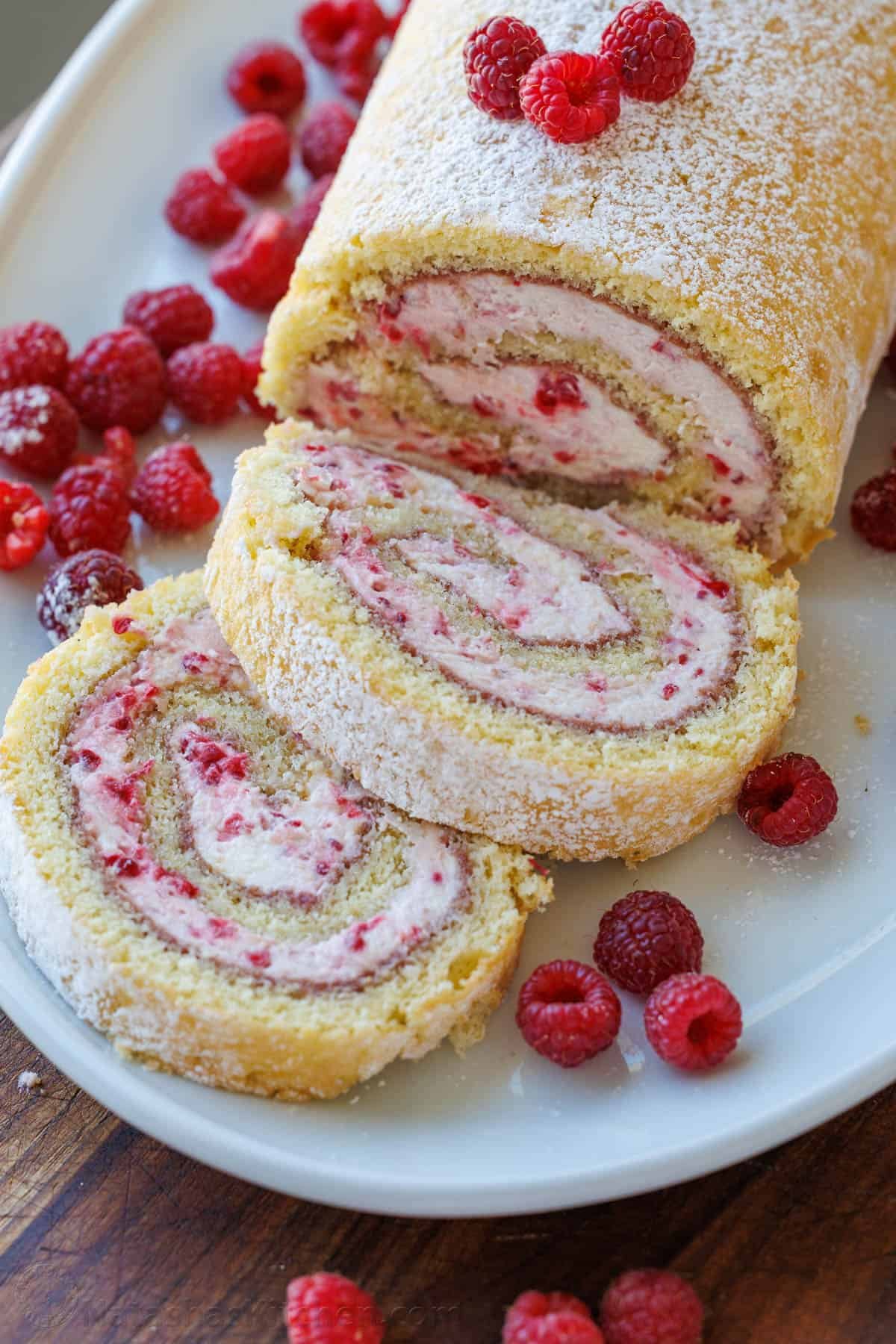 Raspberry Swiss Roll Cake wraps moist sponge cake around a raspberry cream filling. This Raspberry Cake Roll is gorgeous and simple to make. Slices of Raspberry Cake roll on a white plate with raspberries and powdered sugar garnish