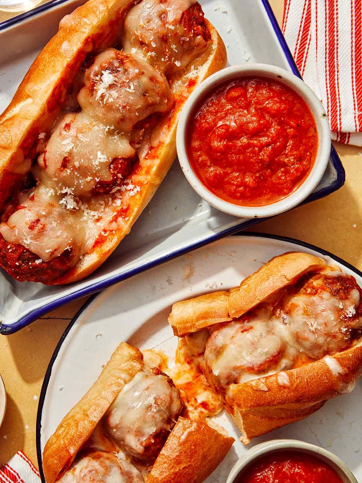Two meatball subs on plates with marinara sauce on the side.