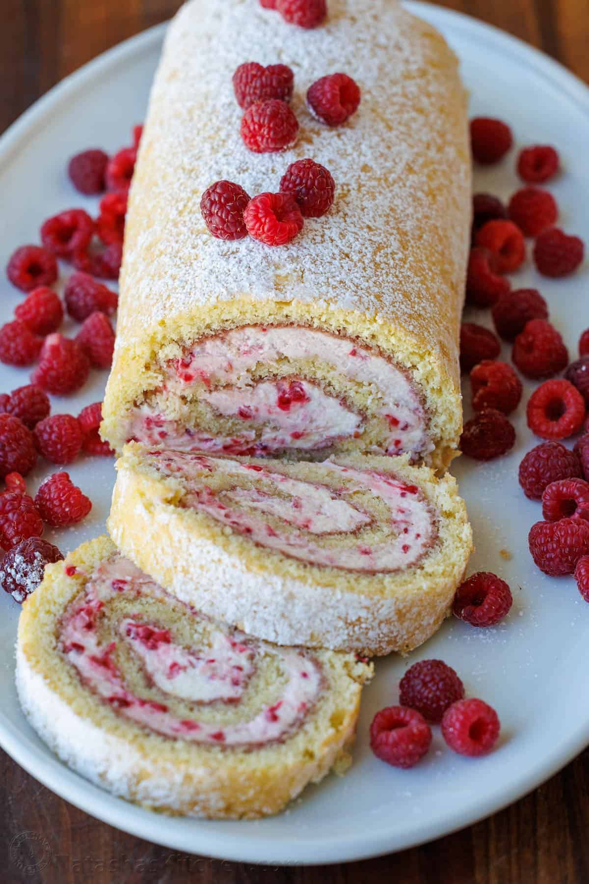 Raspberry Swiss Roll Cake wraps moist sponge cake around a raspberry cream filling. This Raspberry Cake Roll is gorgeous and simple to make. Raspberry Swiss roll cake recipe on a white platter with fresh raspberries scattered around.