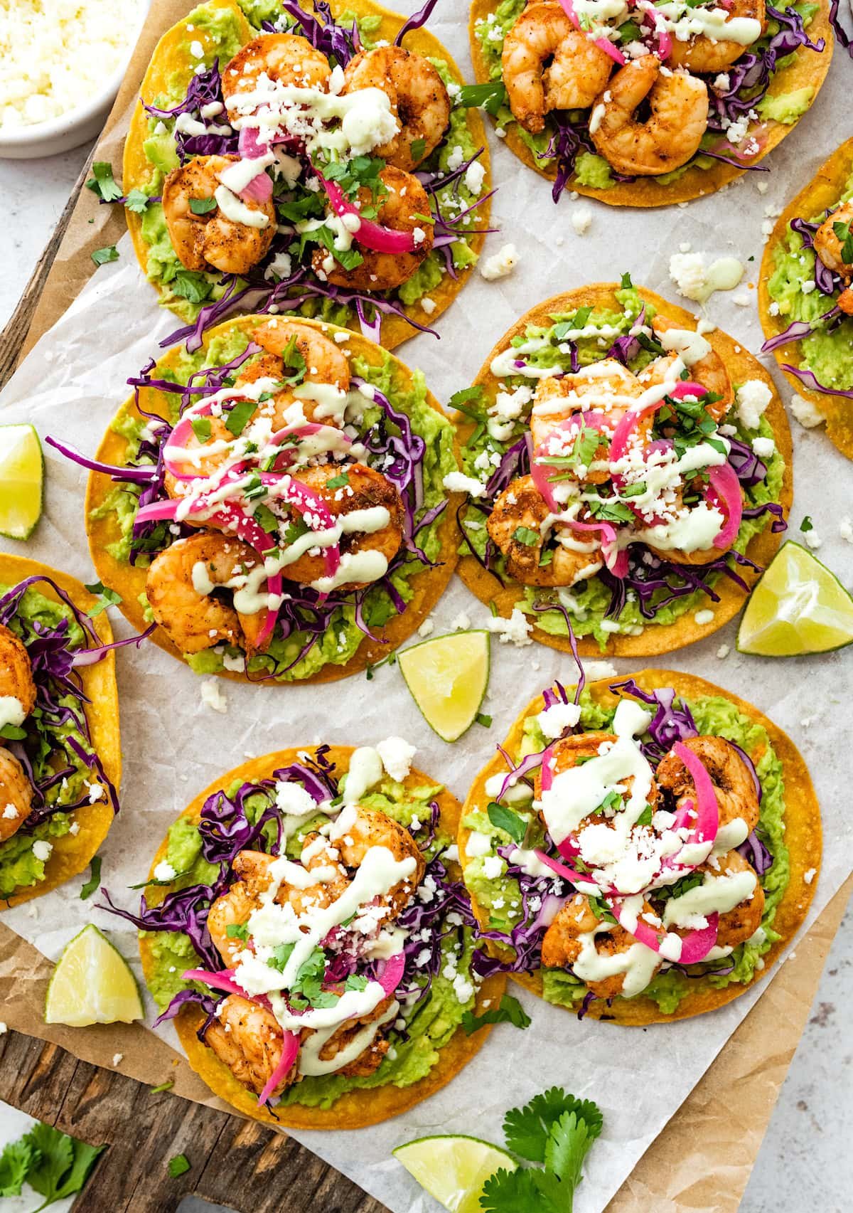 shrimp tostadas topped with avocado, cabbage, pickled red onions, cotija cheese, and jalapeƱo ranch on wood board with parchment paper.