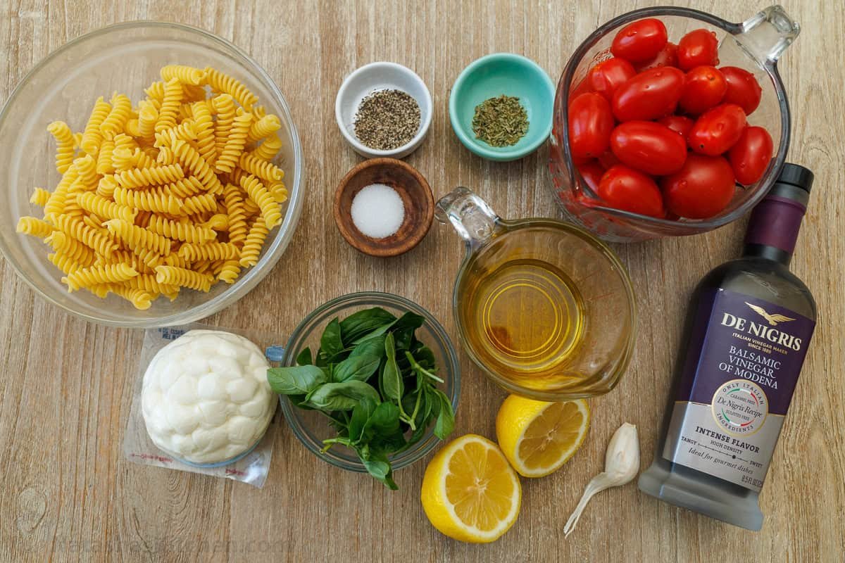 ingredients for Italian side dish recipe including fusilli, mozzarella balls, cherry tomatoes, basil, balsamic glaze, lemon juice, olive oil, salt, pepper, oregano, and garlic cloves