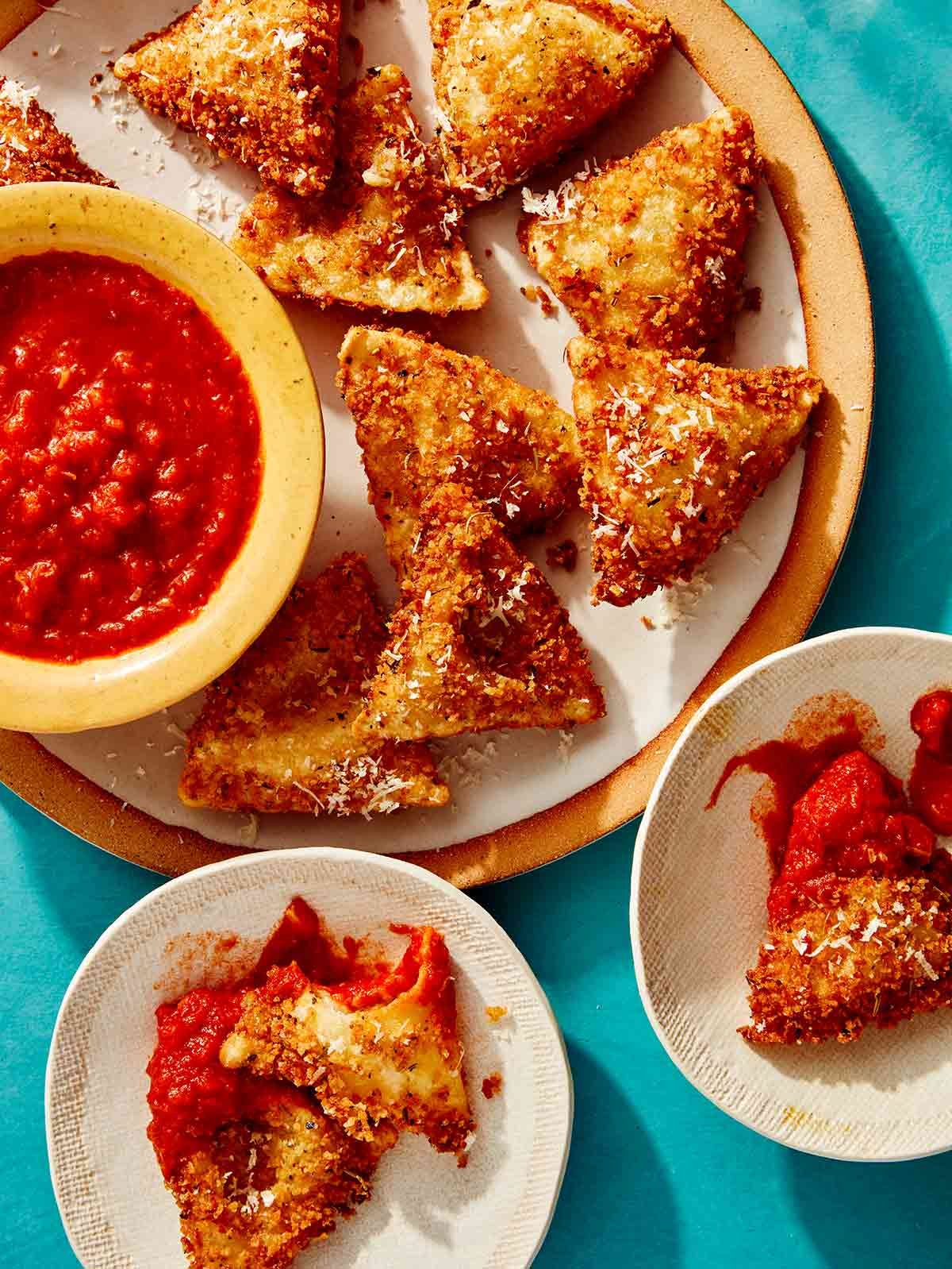 Toasted ravioli on a platter with marinara sauce with some served on plates on the side.