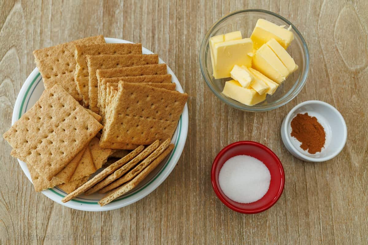 ingredients for making graham cracker crust, including graham crackers, butter, cinnamon and sugar in bowls on a table.
