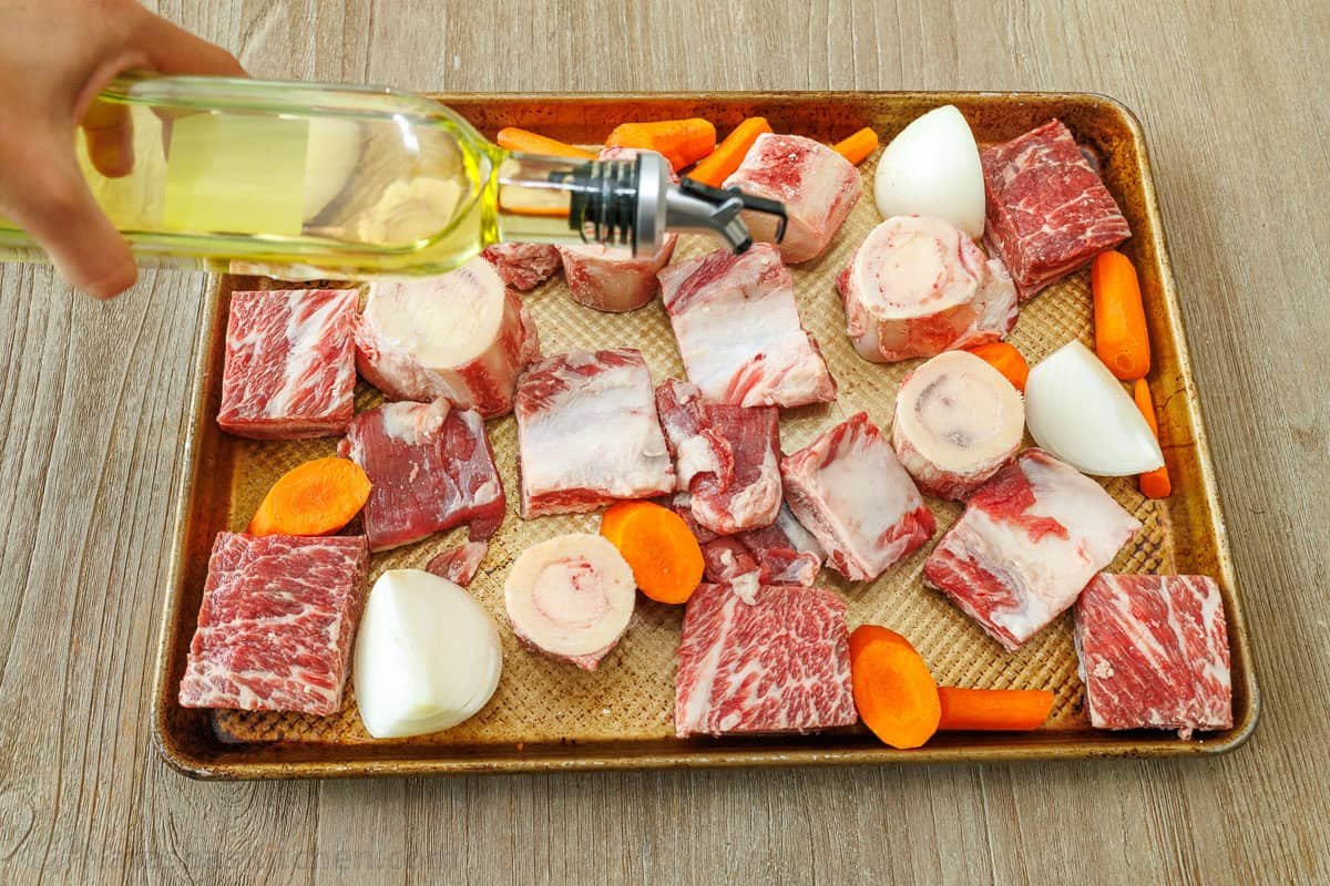 beef bones and vegetables on a roasting pan drizzling with olive oil