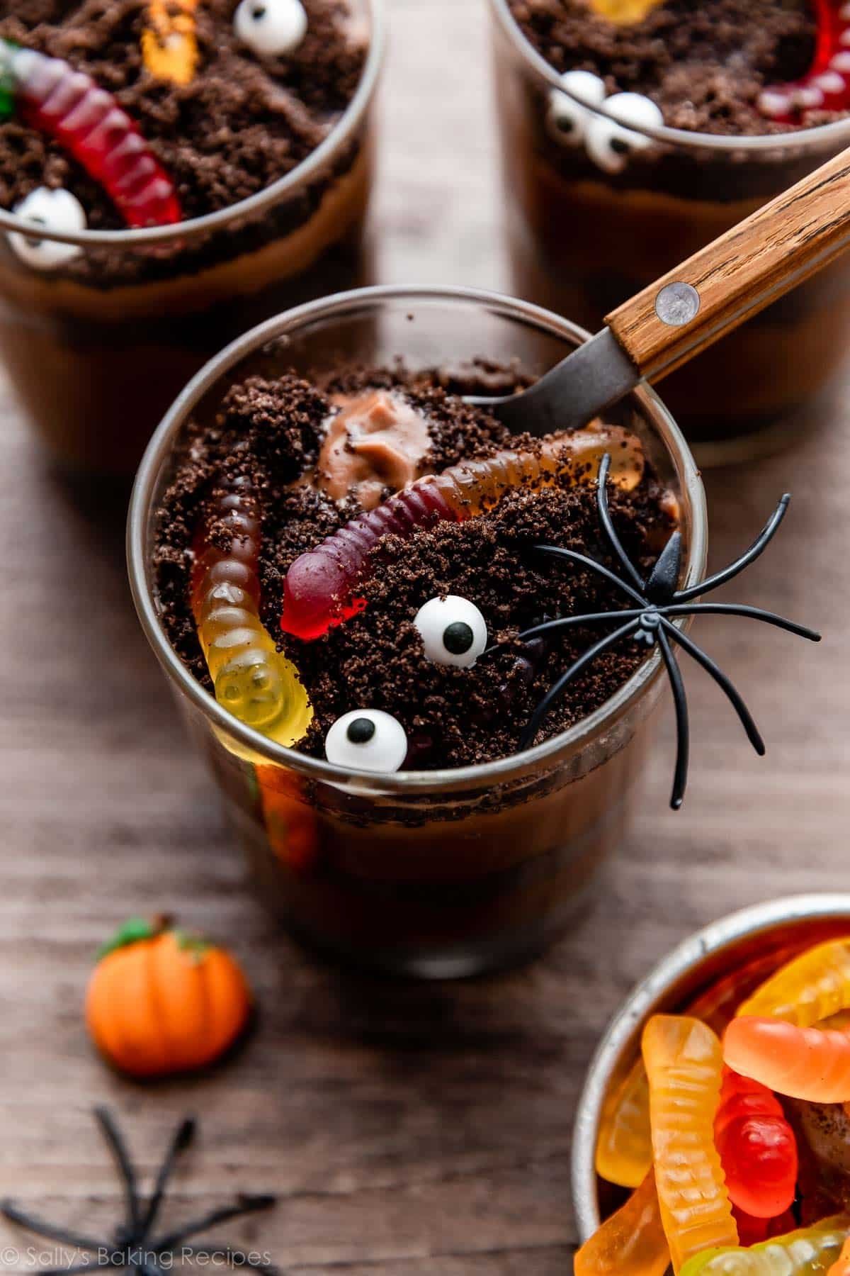 chocolate dirt pudding cups for Halloween with a pretend spider on top, gummy worms buried in crushed Oreos, and a spoon scooping out the 1st bite.