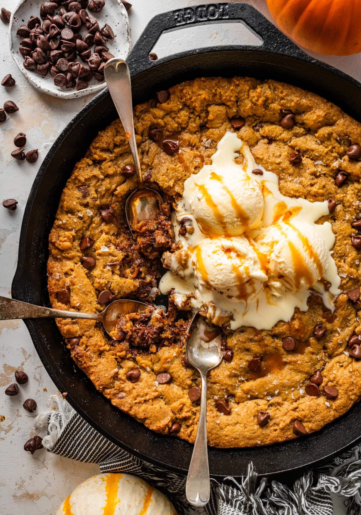 pumpkin chocolate chip skillet cookie with three scoops of vanilla ice cream melting in the center with salted caramel sauce and three spoons digging in.