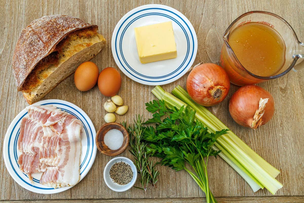 ingredients for the best Thanksgiving side dish with bread, eggs, garlic, butter, onions, celery, parsley, rosemary, salt and pepper, bacon, and chicken stock
