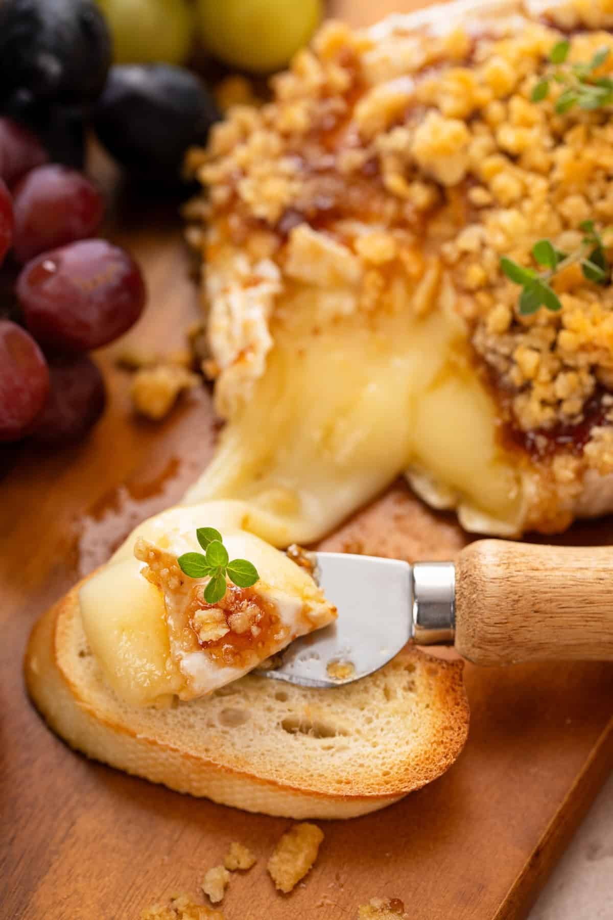 Cheese knife spreading baked brie with jam onto a slice of baguette.