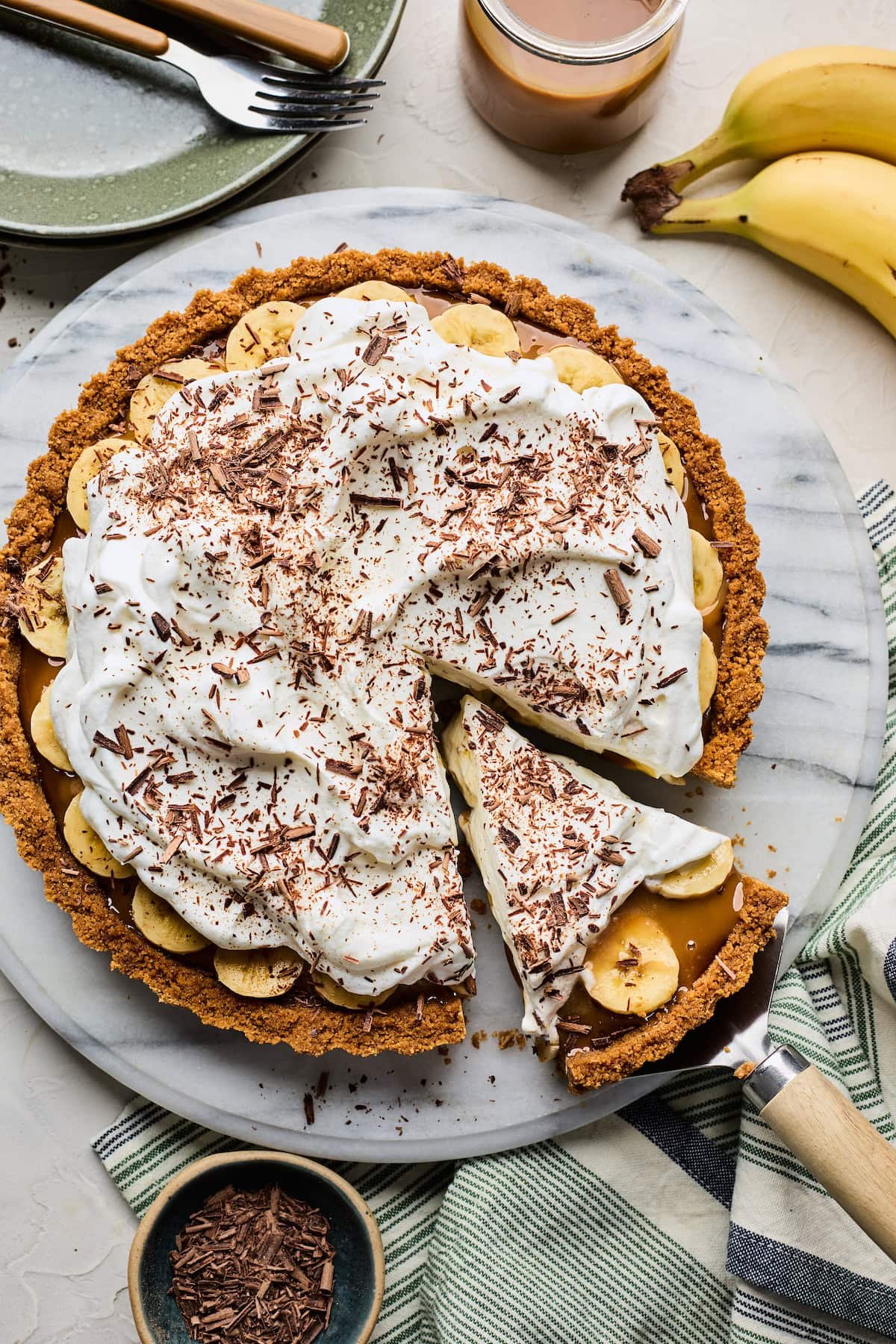 A slice being cut out of Banoffee Pie with whipped cream and chocolate shavings on marble board.