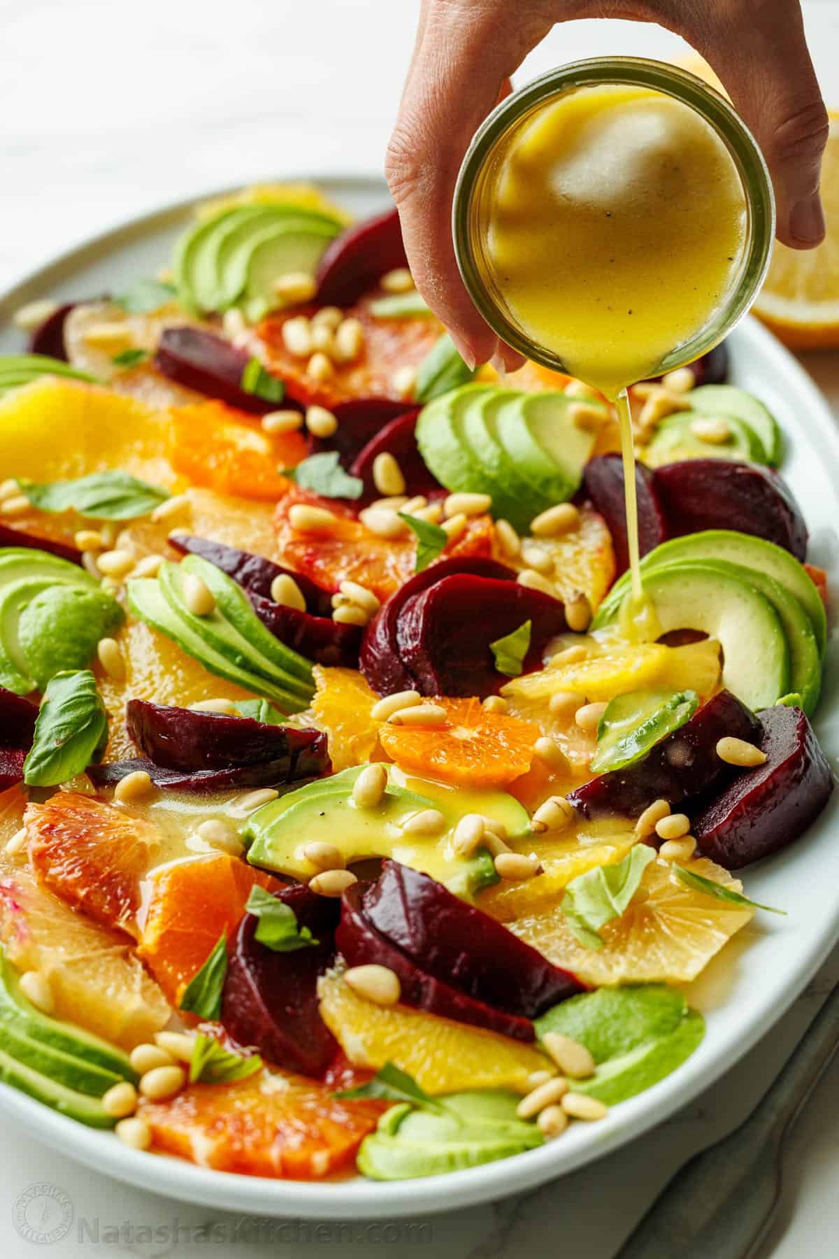 Drizzling lemon vinaigrette over a Citrus salad with fruit and pine nuts on a white platter