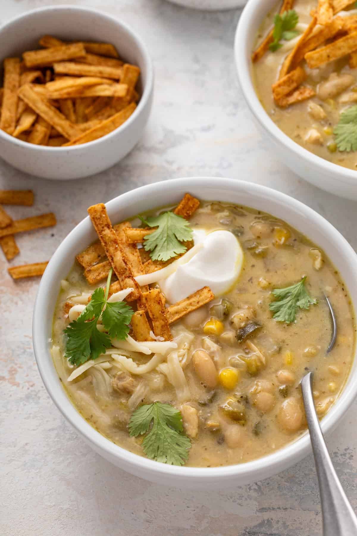Spoon in a bowl of white chicken chili garnished with sour cream, cilantro, and tortilla strips.