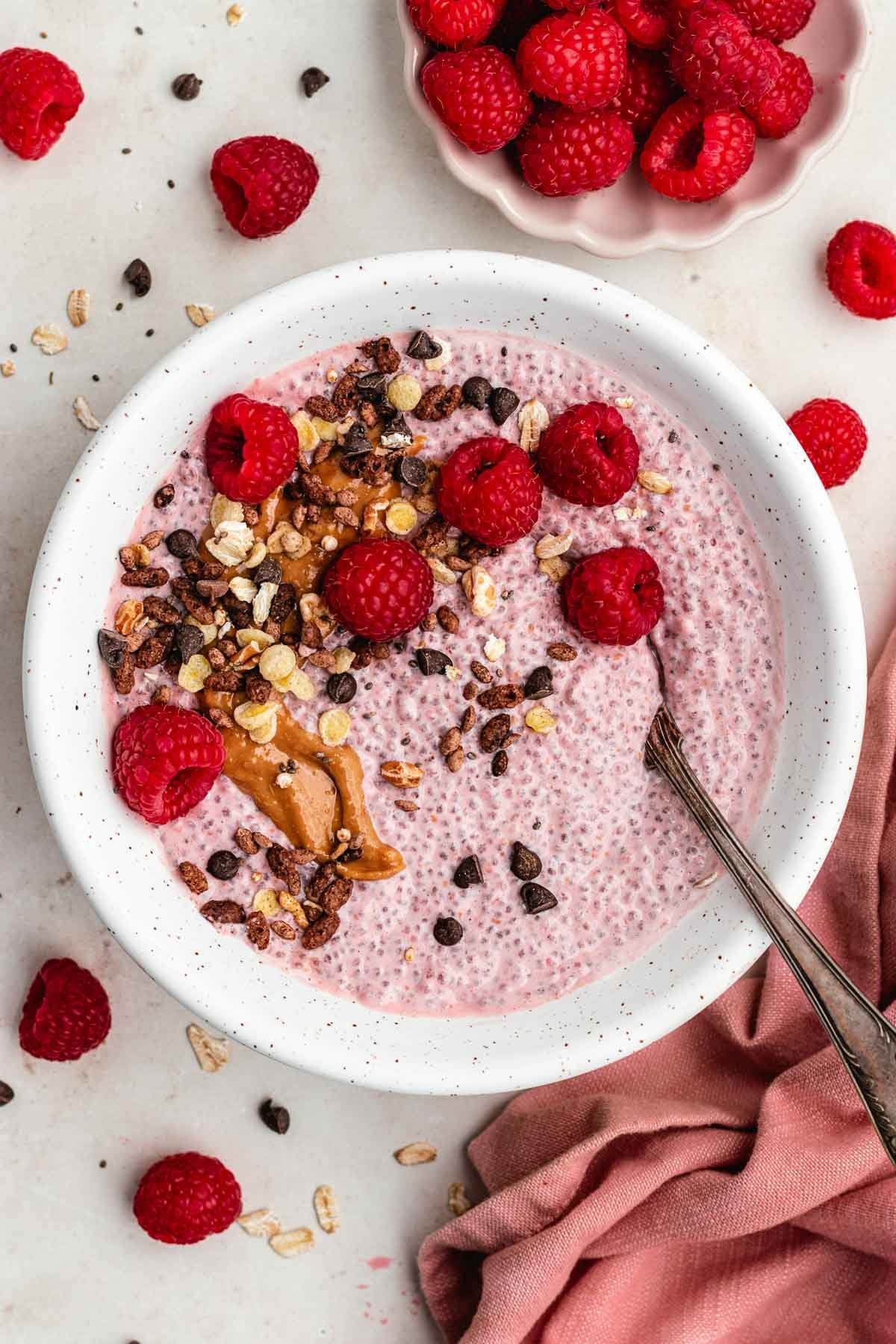 raspberry chia pudding in bowl with a spoon, fresh raspberries, nut butter, chocolate chips, and granola. 