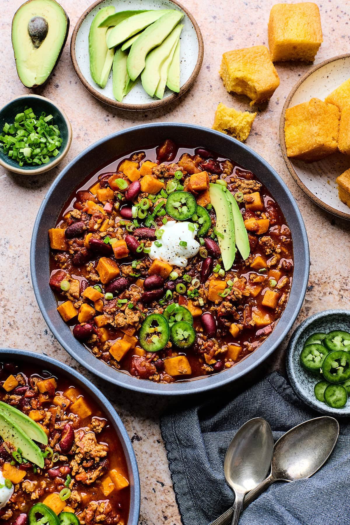 turkey sweet potato chili in bowl with avocado slices, jalapeño slices, and Greek yogurt with a plate of cornbread.