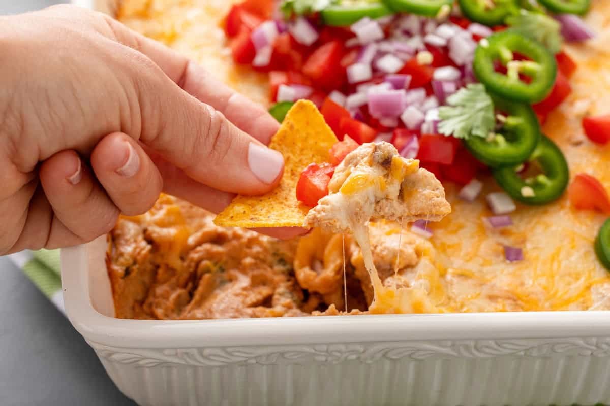 Hand lifting a bite of texas trash dip out of a white baking dish using a tortilla chip.
